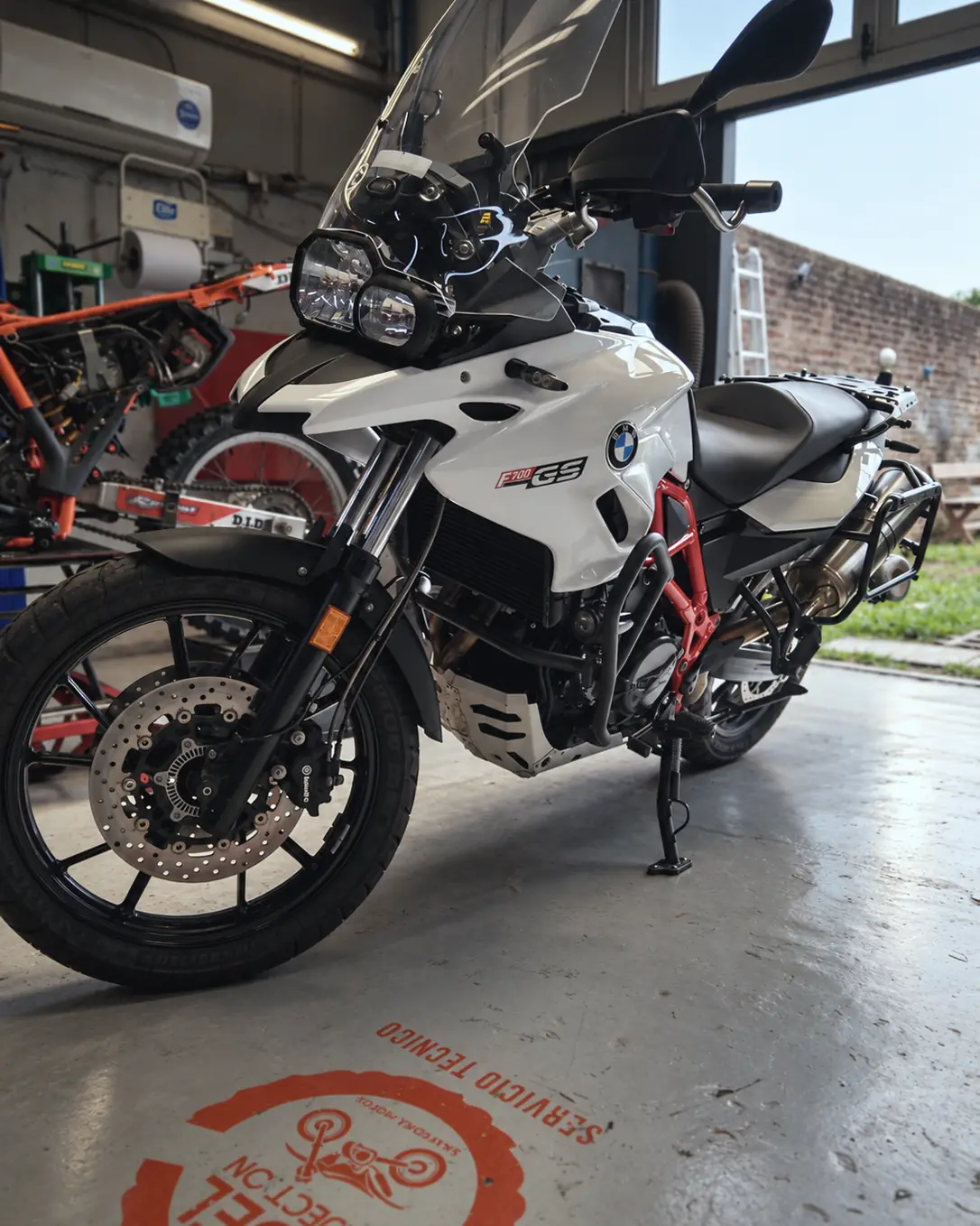 BMW F700 GS blanca en el taller, con el logo de Fuel Injection pintado en el piso.
