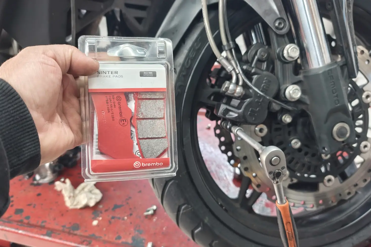 Mano con un blíster de pastillas de freno Brembo nuevas al lado del caliper y el disco delantero de una moto en el elevador, con la llave crique puesta.