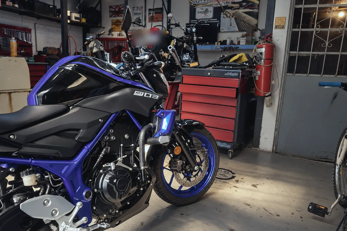 Yamaha MT-03 azul en el taller, con el cartel de inyección electrónica al fondo.
