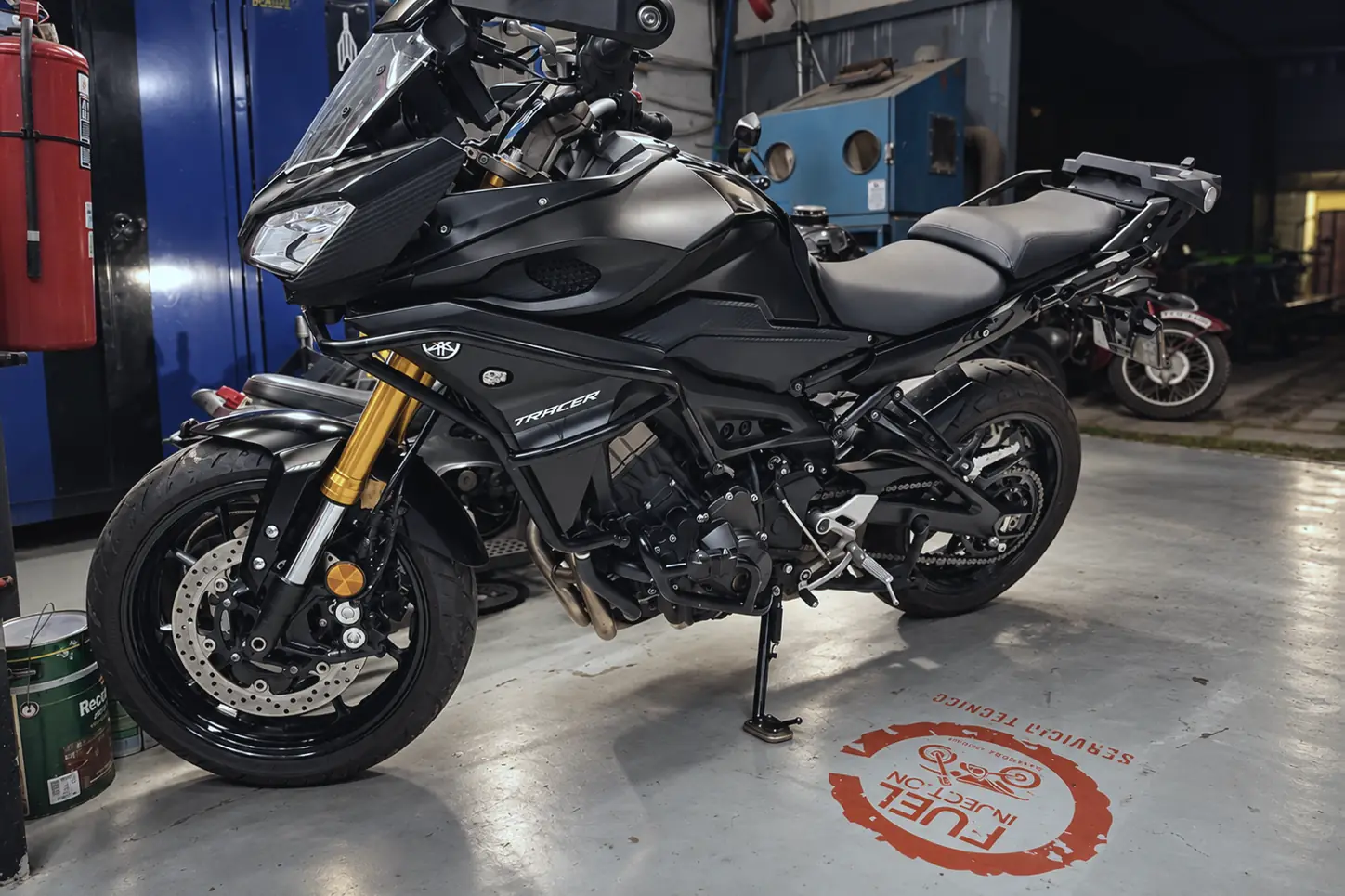 Yamaha Tracer 900 negra en el taller, con el logo de Fuel Injection pintado en el piso.